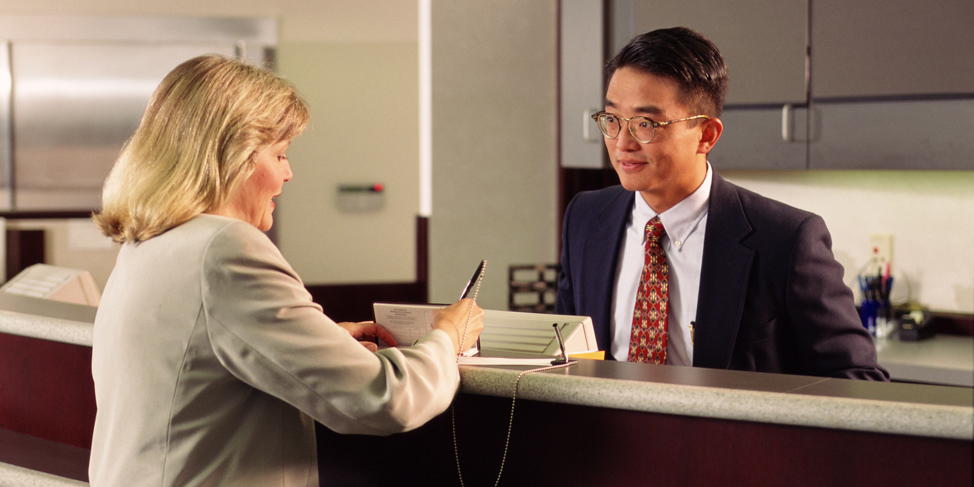  A bank teller wearing a suit and glasses is talking to a customer while working at a counter in a bank on Saturday.