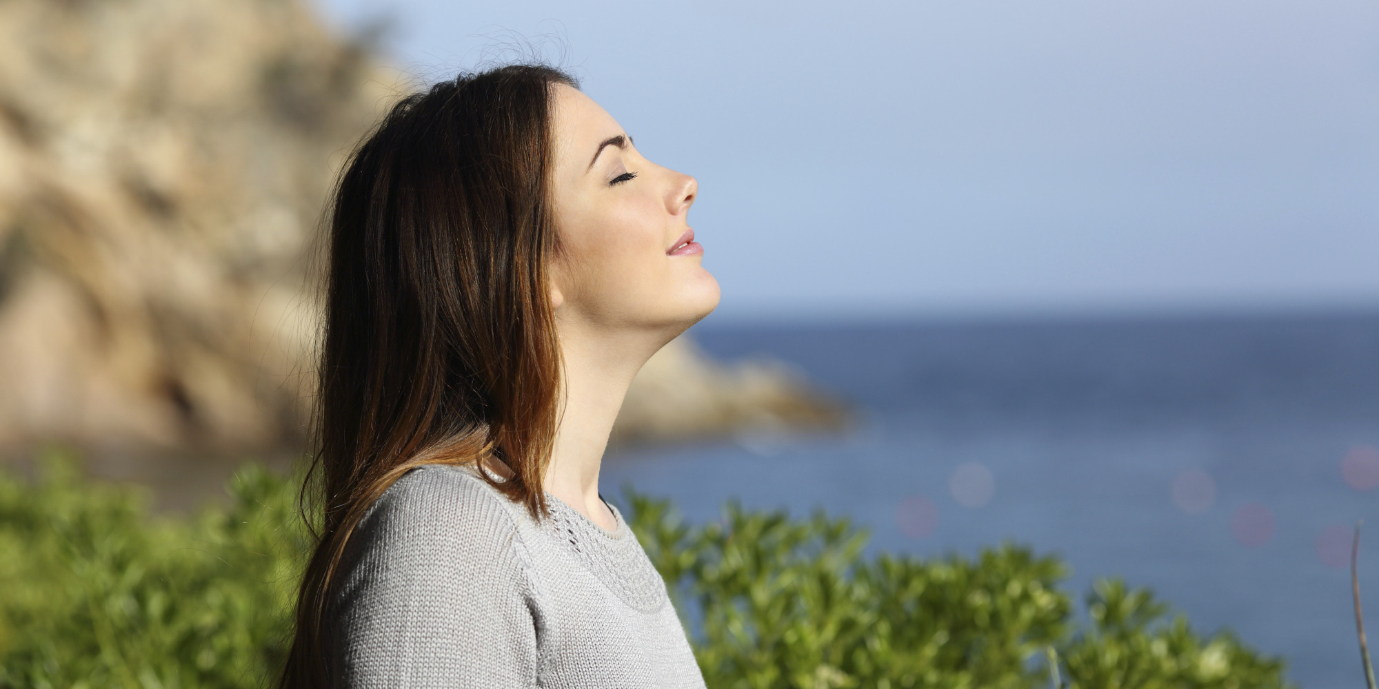  A person with long brown hair sits with their eyes closed and takes a deep breath in the fresh air near the sea.