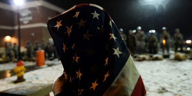 A demonstrator covers himself with a US flag as he joins other to protest outside the police station in Ferguson, Missouri, on November 26, 2014. The streets of Ferguson were decidedly calmer on November 26 after two days of nationwide protests against the decision not to prosecute a white policeman who shot dead an unarmed black teenager. Just a few dozen protesters and clergy braved rain and light snow to protest outside the police department in the St Louis suburb, where 18-year-old Michael B