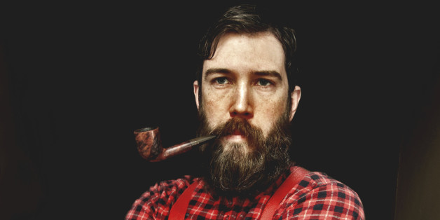 A man dressed as a lumberjack with a plaid shirt and suspenders has a big beard and a pipe in his mouth.