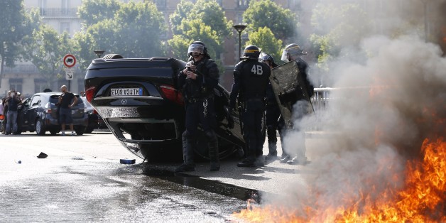 Smoke rises from a fire burning next to French CRS riot police standing near an overturned car as taxi drivers block Porte Maillot in Paris on June 25, 2015. Hundreds of taxi drivers converged on airports and other areas around the capital to demonstrate against UberPOP, a popular taxi app that is facing fierce opposition from traditional cabs. Access to three terminals at Paris-Charles de Gaulle airport and in a number of areas of Paris, especially Porte Maillot, were blocked. AFP PHOTO / THOMAS SAMSON (Photo credit should read THOMAS SAMSON/AFP/Getty Images)
