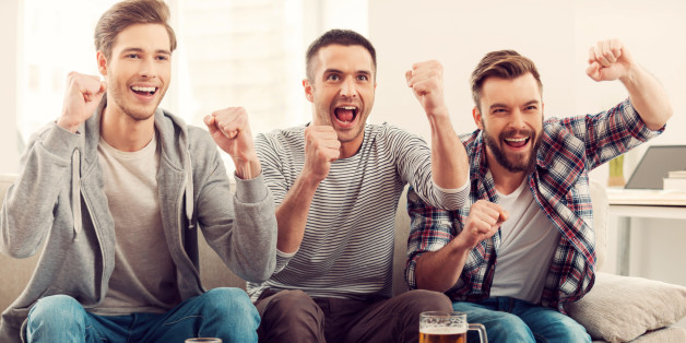 Three happy young men watching football game and keeping arms raised while sitting on sofa