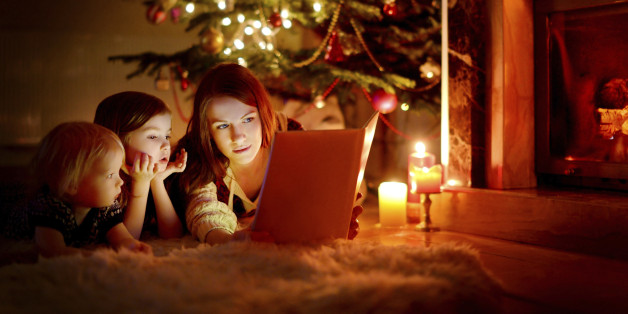 Young mother and her two little daughters reading a book by a Christmas tree in cozy living room in winter