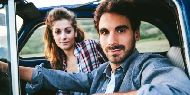 Loving couple in an old blue car. The young man is driving, both look towards the photographer, shot to the inside of the machine. Around the classic landscape of Tuscany, Italy