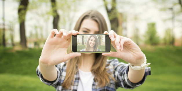 Cute Caucasian brunette teenage woman taking a selfie photo outdoors on sunny summer day in park using her smart phone. Modern youth lifestyle concept.