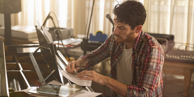A man sitting at a piano writing music, there is a tablet computer with a music app on the piano.