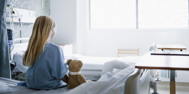 Little girl sitting on a hospital bed with her Teddy Bear by her side looking towards a window.