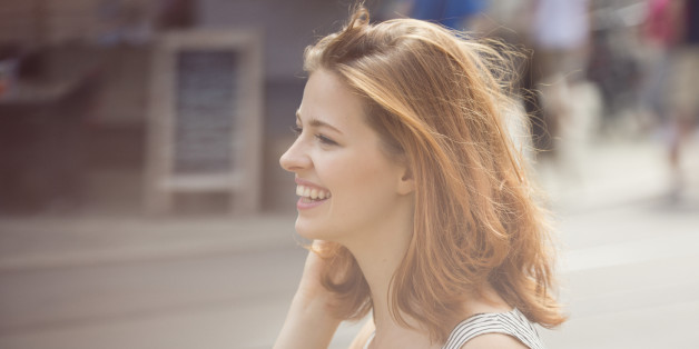 Close portrait of a woman seen from the side. She is laughing and smiling, her hair are flying in the wind as she is walking through the city. She is bathed in a soft sun flare, standing in front of the blurred background of a sunny urban street.