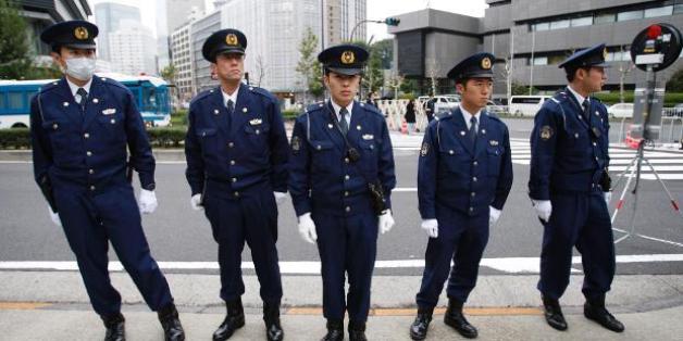 Ein japanischer Polizist riegelt den Tatort ab, im Hintergrund sind Retungswagen zu sehen