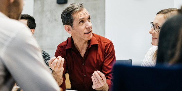A caucasian man is speaking in a business meeting surrounded by colleagues listening to him. He uses his hands and seems convinced and focused.