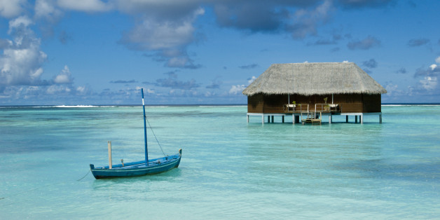 A dhoni (traditional Maldivian boat) floating in a lagoon with an over-water honeymoon bungalow off in the distance, Maldives.