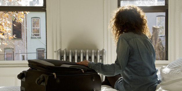 Female solo traveller sat on bed with suitcase looking out of the window in Brooklyn, New York.