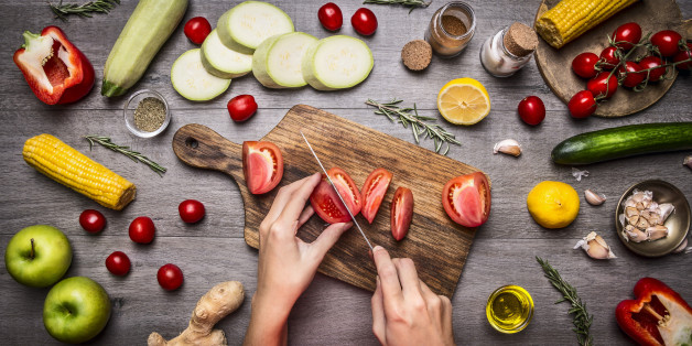 Female hand cut tomatoes on rustic kitchen table, around lie ingredients, vegetables, fruits, and spices, Healthy foods, cooking and vegetarian concept.
