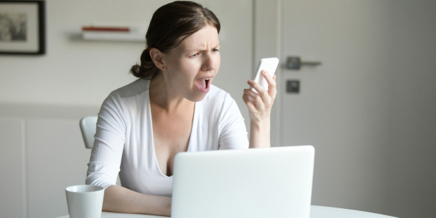 Portrait of a young woman at the desk with a laptop, looking questionnaire at the screen of the mobile she holds at her hand. Business concept photo, lifestyle