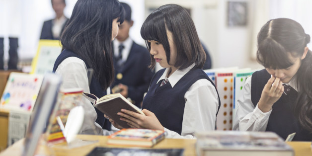 High school girl reading book in classroom. Female student is with classmates in brightly lit school. They are in uniforms.