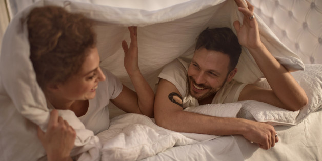 Young happy couple lying in bed and having fun while being covered with a duvet. Focus is on happy man.