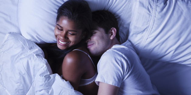 Photo of an attractive young mixed race couple, lying in bed as husband playfully invades his wife's side of the bed.