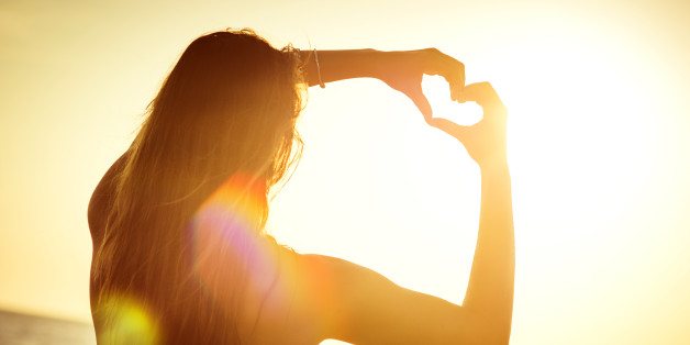 Young woman at the beach, making the shape of a heart with her hands, towards the sun.