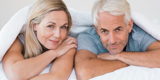 Senior romantic couple under a white duvet in the bed. Happy mature couple lying down in their bed and looking at camera. Retired man and smiling woman having fun lying on bed at home.