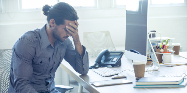 Stressed business man at the office. He is casually dressed and looking distraught. He looks very uncomfortable and could also have a headache. He is has his head in his hand and looks very upset. Hi is sitting at a desk with a computer and phone. Copy space.