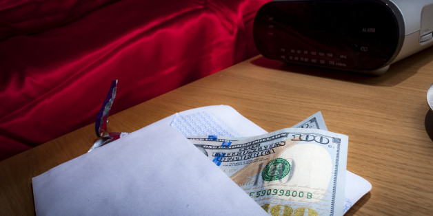 Prostitution concept with money in an envelope on a nightstand in a hotel room, representing the payment for a prostitute, escort or sex worker, next to the bed covered in red satin sheets