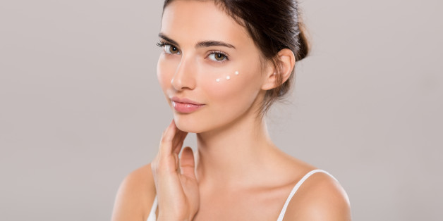 Young woman applying moisturizer on face isolated on grey background. Beautiful woman applying cosmetic cream on skin near eyes and looking at camera. Beauty and skin care concept.