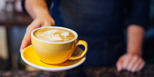 A barista serves up a hot espresso latte in a classy ceramic mug with heart shaped fern poured into the steamed milk foam. Vertical. Extremely shallow depth of field with selective focus on barista's finger.