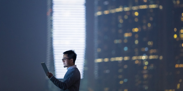 Smart businessman using and looking at digital tablet in city, standing against illuminated financial skyscrapers at night time.