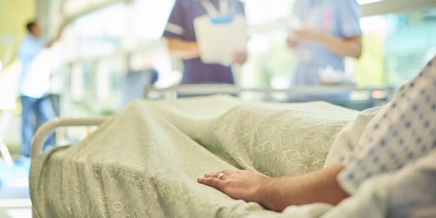 a female senior nurse or matron is chatting to a young a young student nurse is attending to the dressings trolley at the foot of a patient's bed . They are all defocussed apart from the patient's hand in the foreground.
