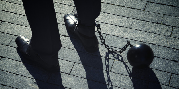 'Long, dark shadows hang from a the feet of a businessman standing with iron ball and chain'