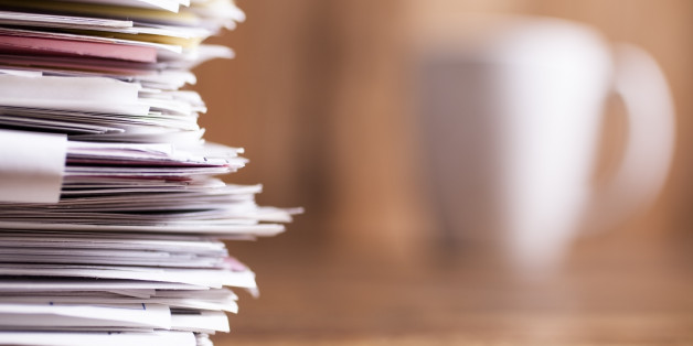 Large stack of multi-colored files and paperwork lie on an office desk. A white coffee mug in background. Wooden backdrop. Copyspace to side. No people.