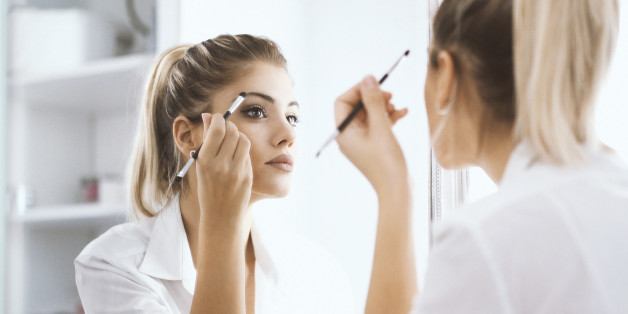 Closeup rear view of attractive mid 20's blond woman putting on some makeup before heading to work. She's in front of large bathroom mirror, focus is on her reflection.
