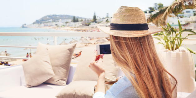 Happy young blond woman taking smartphone photo with smart phone of a beach in Gammarth Tunis, Tunisia on outdoor patio sofa furniture