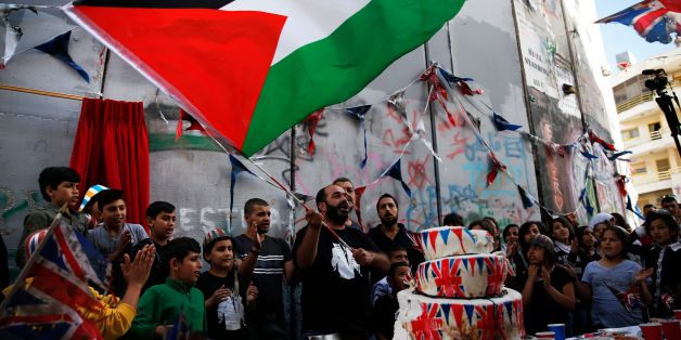 A Palestinian man flies his national flag above a cake bearing images of the Union Jack as children from the al-Aida refugee camp attend an event held by secretive British street artist Banksy to apologise for the 100th anniversary of the Balfour Declaration on November 1, 2017 at his Walled-Off Hotel in Bethlehem in the occupied West Bank.The queen revealed a plaque carved in stone saying 'Er, Sorry,' playing on the common initials for Elizabeth Royal. / AFP PHOTO / AHMAD GHARABLI (Photo