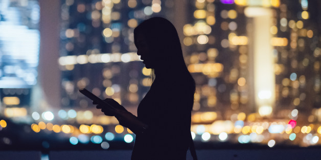 Silhouette of woman using digital tablet device in city at night, with illuminated and blurry commercial skyscrapers in the background.