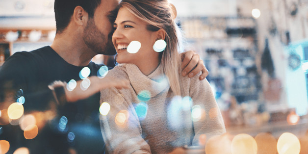 Closeup of a mid 20's couple spending romantic evening at a cozy coffee place and having some fun. He's whispering someting sweet into her ear and that made her smile.Shot from the outside through the large window they are sitting next to. Lots of defocused glimmering lights in the frame.