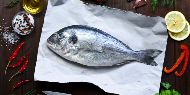 Top view of a raw sea bream fish on paper at the center of an horizontal frame with cooking and seasoning ingredients like olive oil, salt and pepper, rosemary, lemon, thyme, chili pepper, tomatoes, parsley, garlic, chive, are all around the fish. DSRL studio photo taken with Canon EOS 5D Mk II and Canon EF 70-200mm f/2.8L IS II USM Telephoto Zoom Lens