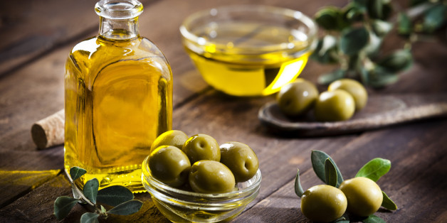 Horizontal shot of an olive oil bottle and a little glass bowl filled with green olives on rustic wood table. Two olives with leaves are at the bottom-right while a bowl filled with olive oil is out of focus at background beside olive tree branch. Predominant colors are gold, green and brown. DSRL studio photo taken with Canon EOS 5D Mk II and Canon EF 70-200mm f/2.8L IS II USM Telephoto Zoom Lens